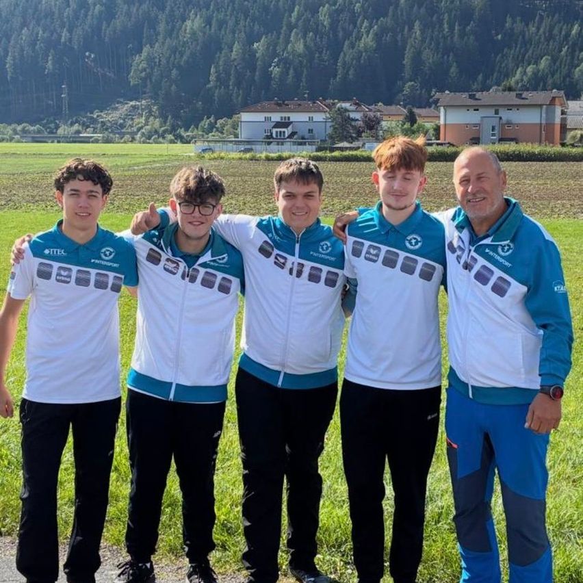 Fünf Personen stehen in einer Reihe und lächeln für ein Foto. Sie tragen passende weiße und blaue Sportjacken. Dahinter befindet sich ein Grasfeld mit einem Haus und Bäumen in der Ferne.