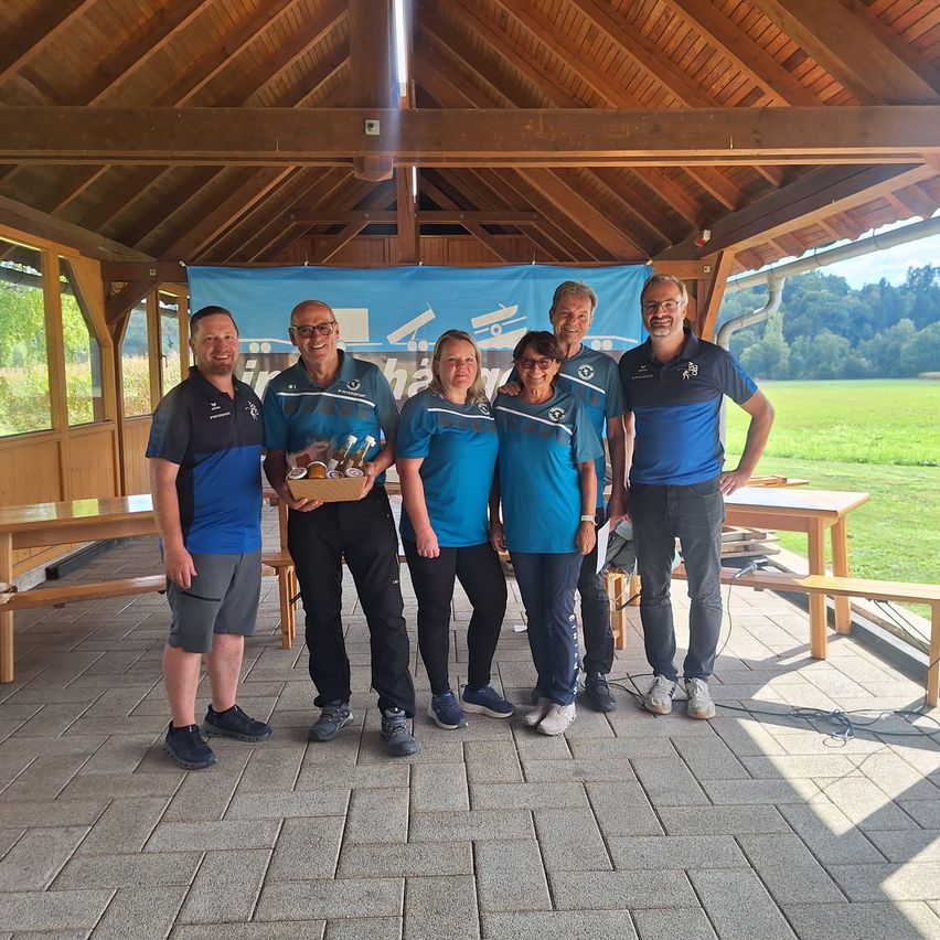 Eine Gruppe von Menschen in passenden blauen Shirts posiert für ein Foto unter einem hölzernen Pavillon. Sie halten einen Korb mit Gegenständen.