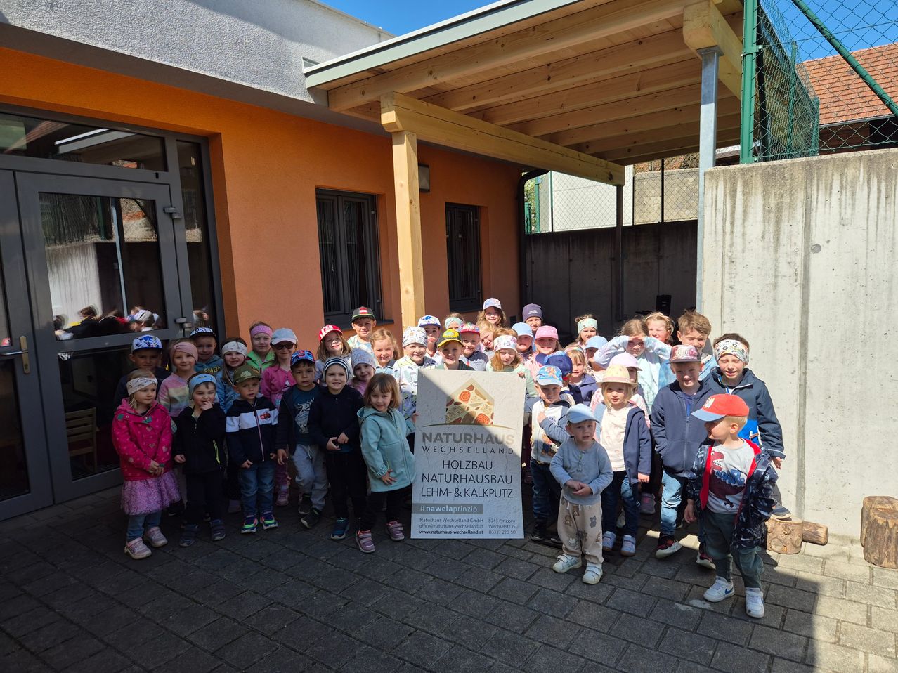 Eine Gruppe von Kindern und Erwachsenen posiert für ein Foto vor dem Holzbau Naturhaus in Wechselland, Österreich. Die Gruppe steht vor einem Gebäude mit einer Holzveranda. Einige Leute tragen Hüte und Turnschuhe. Ein Plakat mit dem Text 'Naturhaus Wechselland Holzbau Lehm & Kalkputz' steht in der Mitte der Gruppe.