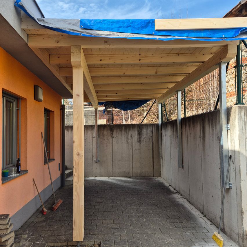 Eine offene Terrasse unter einem hölzernen Vordach. Ein blauer Tarp bedeckt das Dach. Ein Besen und eine Kehrschaufel stehen neben einem Haufen Ziegel.
