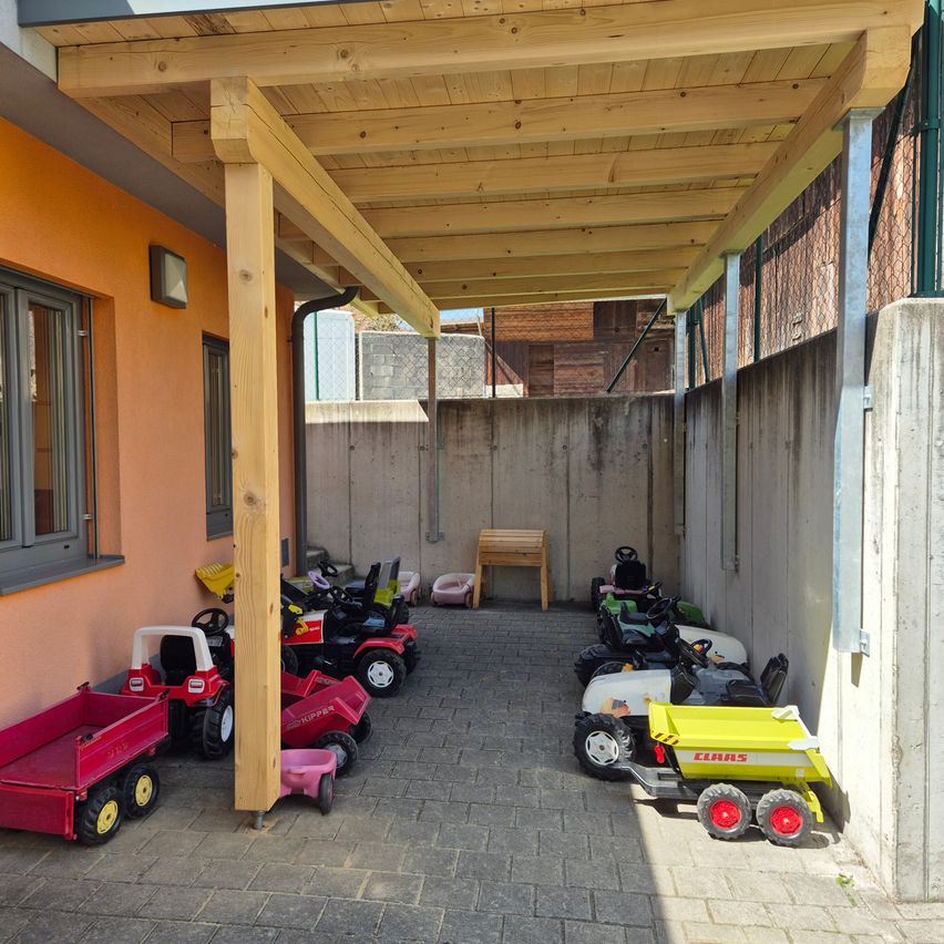 Ein überdachter Außenbereich mit Ziegelboden. Einige Spielzeugautos sind in einer Reihe geparkt und einige an der Wand. Eine Holzbank steht rechts.
