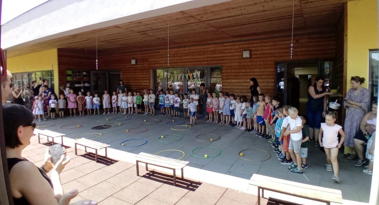 Eine Gruppe von Kindern und Erwachsenen steht in einer Reihe in einem Raum mit Holzwänden und einem Schild, das 'Sommerfest' steht. Sie sind von Hula-Hoops umgeben.