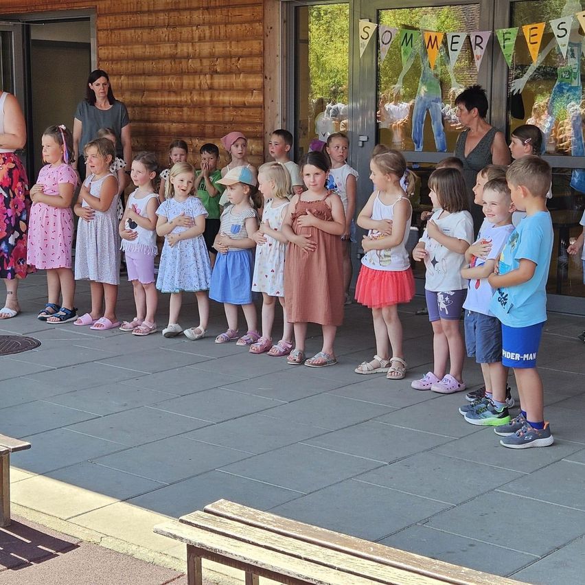 Kinder in Sommerkleidung stehen vor einem Gebäude mit Glastüren, an denen Wimpel wehen. Erwachsene sind anwesend, und die Hände der Kinder sind vor ihnen verschränkt.