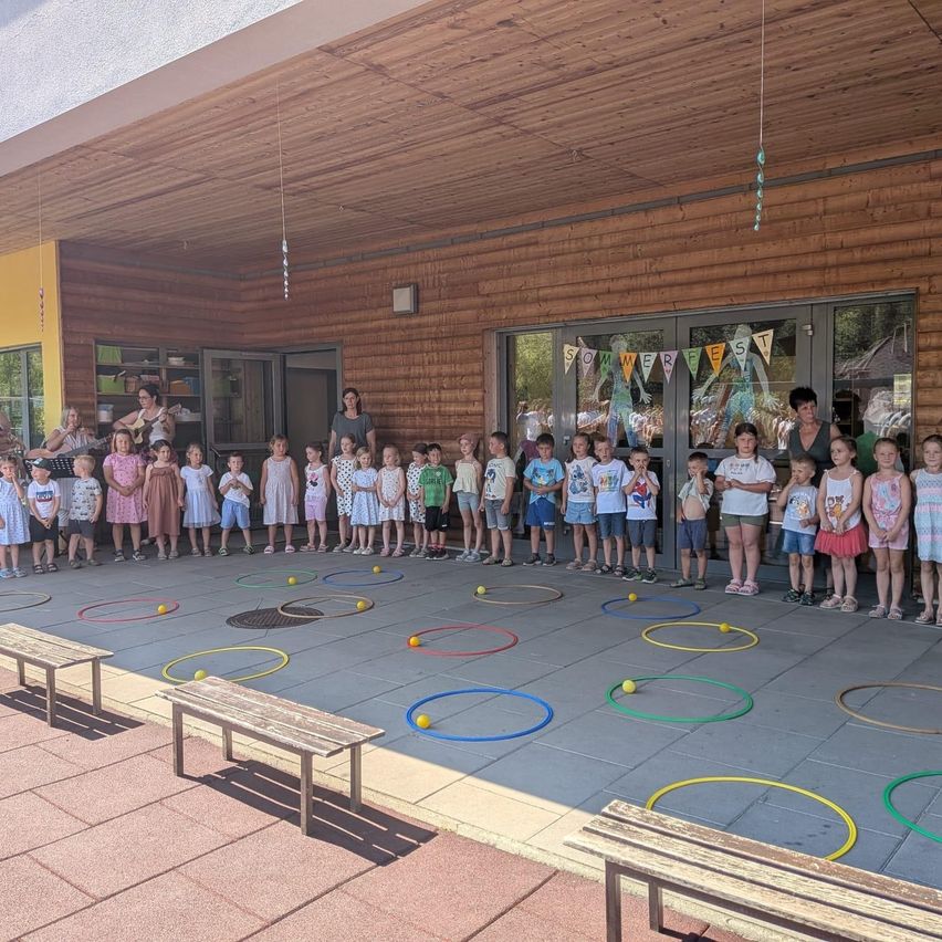 Eine Gruppe von Kindern und Erwachsenen steht in einem Kreis um einen Innenhof mit bunten Hula-Hoops. Zwei Frauen spielen Gitarre und singen.