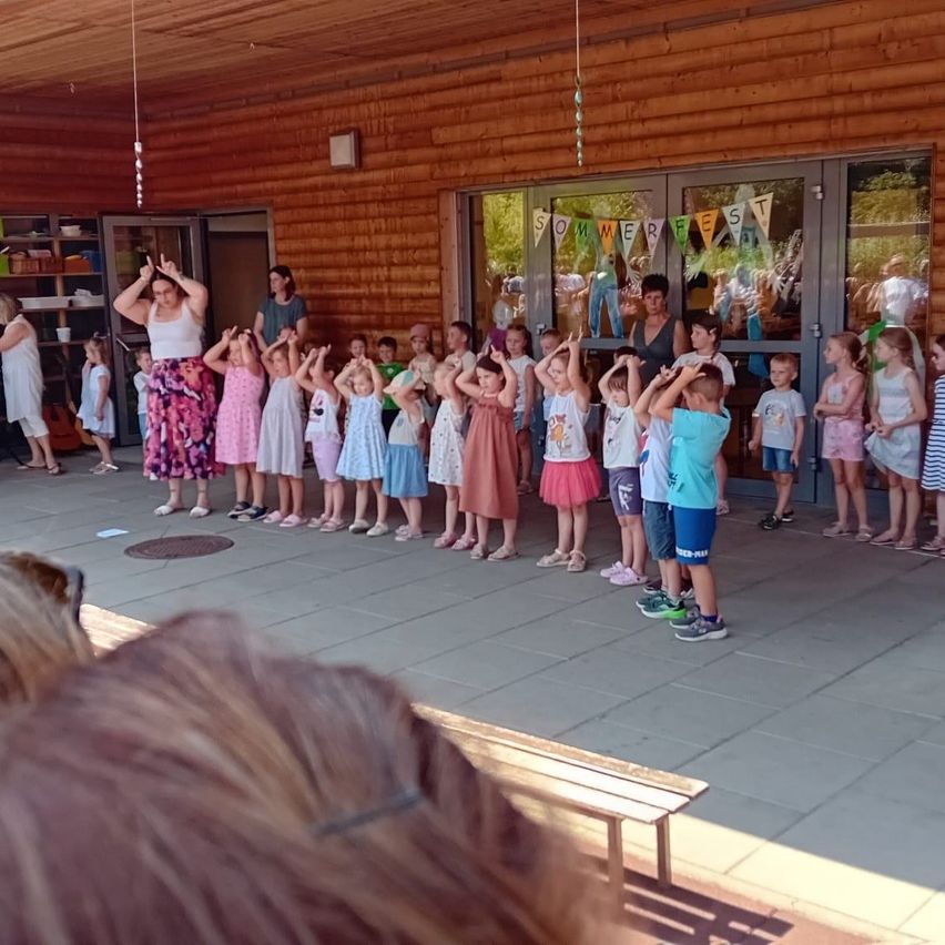Eine Gruppe von Kindern und Erwachsenen versammelt sich in einem Raum mit Holzwänden. Die Kinder stehen in einer Reihe und heben ihre Hände. Eine Frau in einem weißen Tanktop führt die Gruppe an. Über dem Eingang befindet sich ein Banner mit der Aufschrift 'Sommerfest'.