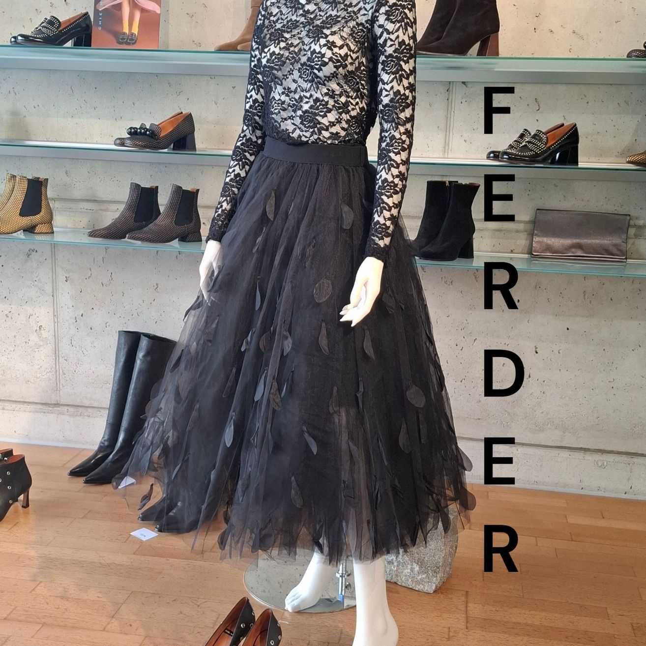 Ein schwarzes Kleid mit Spitzenärmeln und einem Tutu-Rock wird auf einem Mannequin in einem Bekleidungsgeschäft ausgestellt, mit Regalen verschiedener Schuhe.
