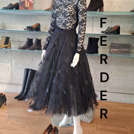 Ein schwarzes Kleid mit Spitzenärmeln und einem Tutu-Rock wird auf einem Mannequin in einem Bekleidungsgeschäft ausgestellt, mit Regalen verschiedener Schuhe.