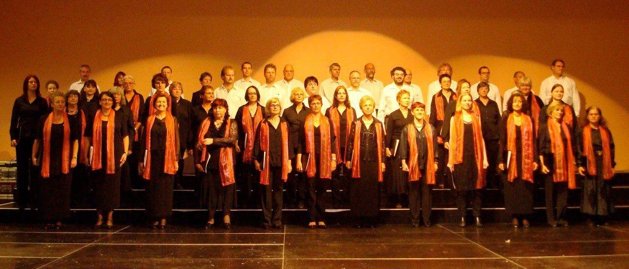 Eine Gruppe von Menschen in schwarzen und orangefarbenen Schärpen steht auf der Bühne, wahrscheinlich bei den World Choir Games in Graz 2008. Ein Banner hinter ihnen trägt die Aufschrift '5. World Choir Games Graz 2008.'