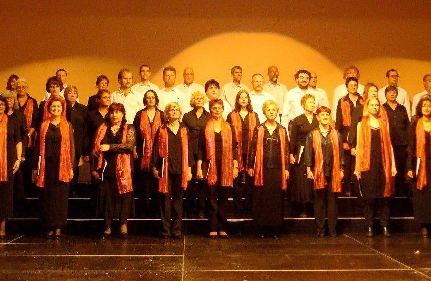 Eine Gruppe von Menschen in schwarzen und orangefarbenen Schärpen steht auf der Bühne, wahrscheinlich bei den World Choir Games in Graz 2008. Ein Banner hinter ihnen trägt die Aufschrift '5. World Choir Games Graz 2008.'