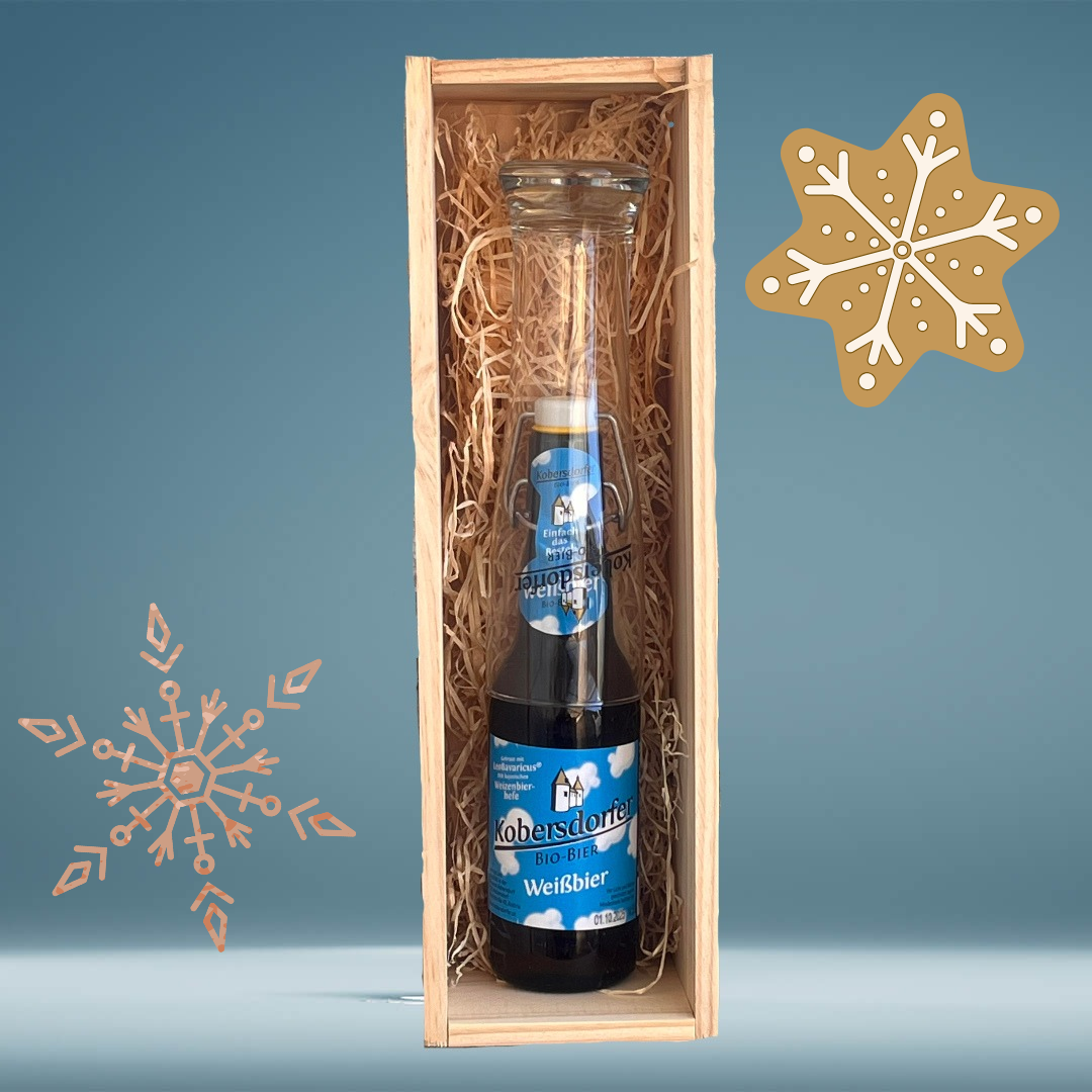 Eine Holzkiste enthält eine Flasche Koebersdorfer Weißbier. Die Flasche ist dunkel und hat ein blaues Etikett. Die Kiste ist mit Stroh gefüllt. Schneeflocken sind auf dem blauen Hintergrund gezeichnet.