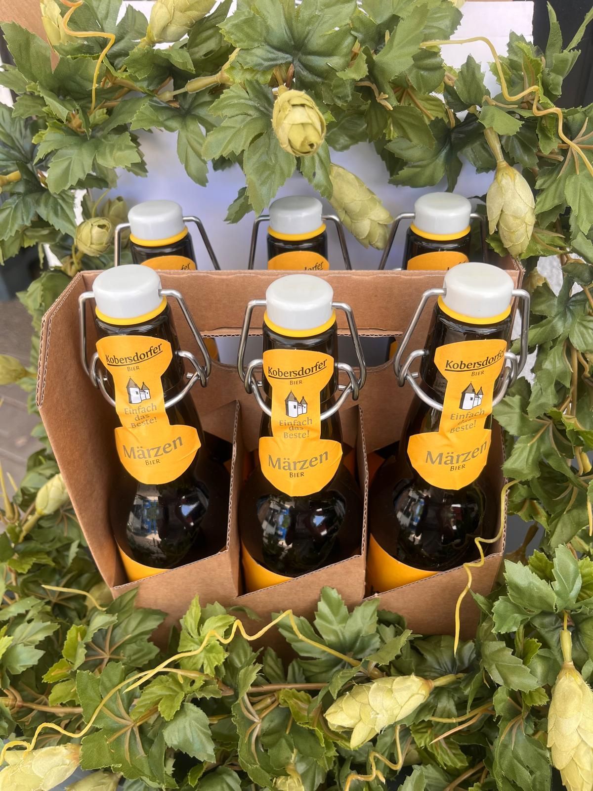 Eine Schachtel mit sechs Flaschen Maerzen Bier, mit weißen Kappen und gelben Etiketten mit einem Burglogo. Die Flaschen werden von Metallklemmen gehalten und sind von grünen Blättern umgeben.