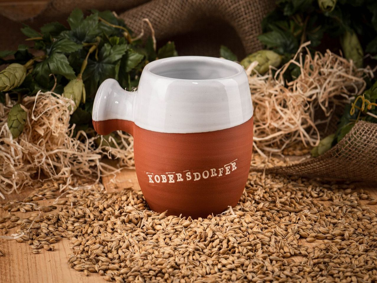 Eine Ton-Tasse mit weißem Henkel und braunem Boden, beschriftet mit Kobersdorfer, steht auf einem Haufen Getreide.