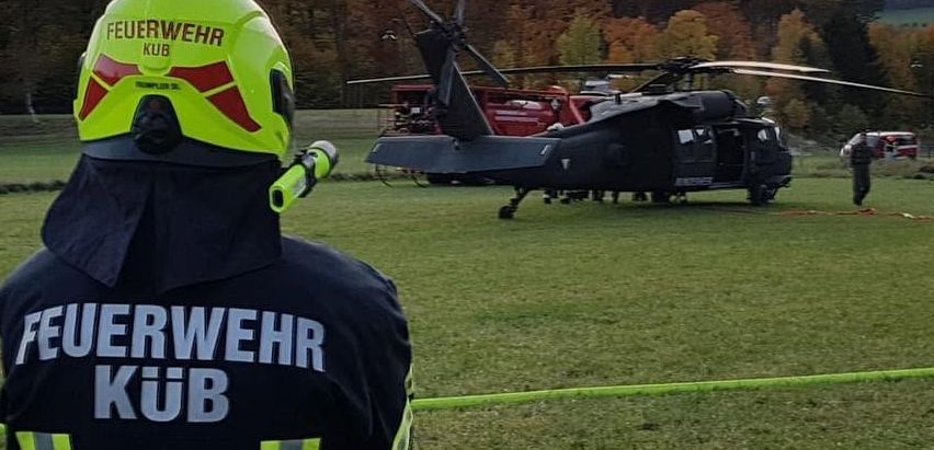 Ein Feuerwehrmann in einem gelben Helm steht auf einem Rasenfeld mit einem Hubschrauber im Hintergrund.