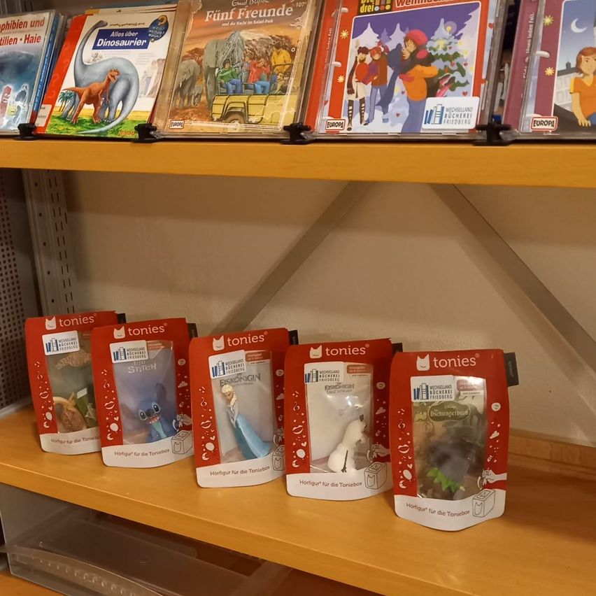 Ein hölzerne Regal mit fünf Tonies-Spielzeugfiguren in roter Verpackung ist ausgestellt. Die Figuren sind mit ihren Charakternamen beschriftet. Über den Spielzeugen befinden sich Bücher mit Illustrationen und Text. Der Hintergrund ist eine weiße Wand.