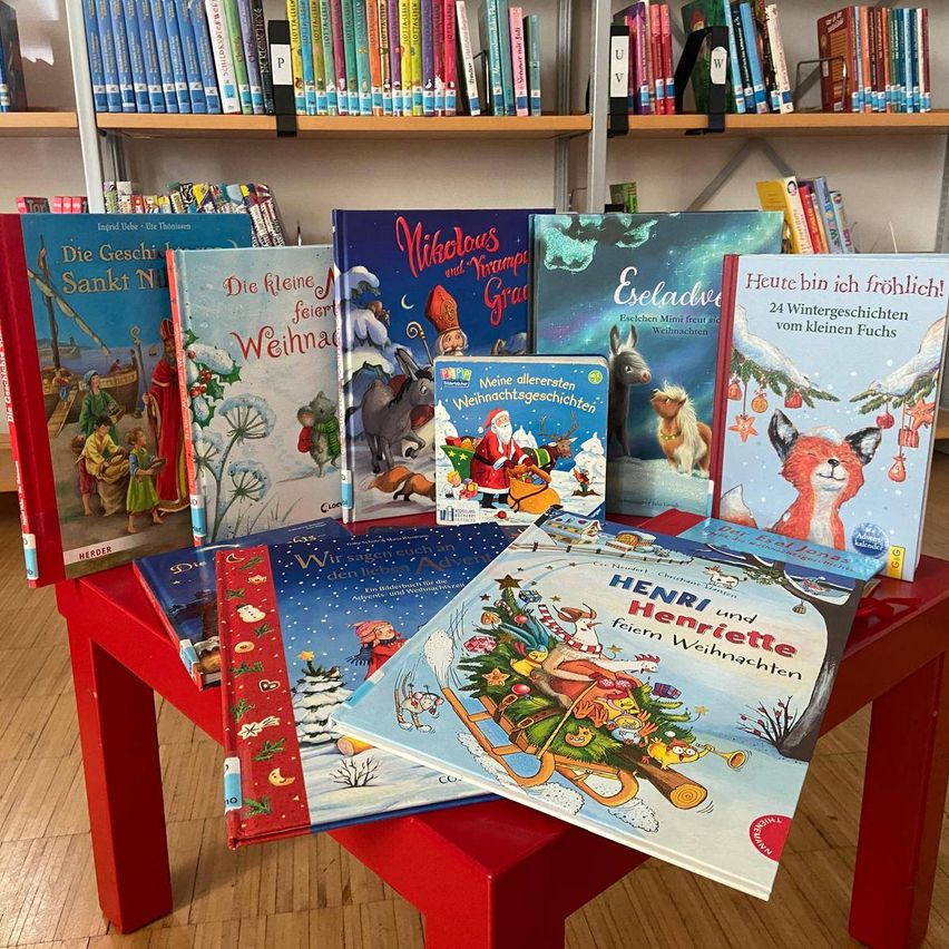 Mehrere Weihnachtskinderbücher sind auf einem roten Tisch ausgestellt. Dahinter ist ein Bücherregal mit verschiedenen Büchern zu sehen.