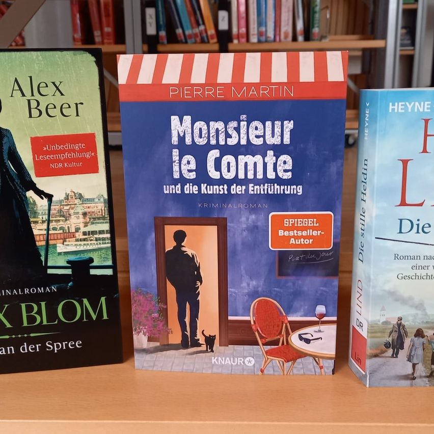 Three books on a wooden shelf: 'Alex Beer', 'Monsieur le Comte', and 'Die stille Heldin'. 'Monsieur le Comte' is a blue book with an illustration of a man entering a room.