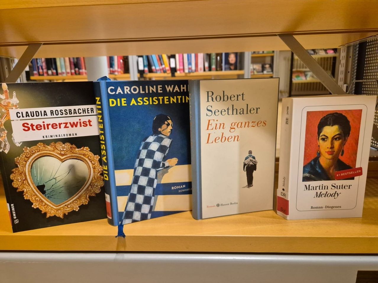 A wooden shelf in a library holds four books. The titles are Caroline Wahr, Die Assistentin, Robert Seethaler, Ein ganzes Leben, Rossbacher, and Martin Meloty.