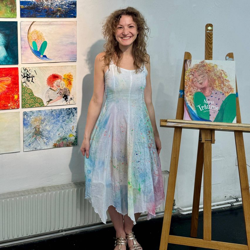 Eine Frau steht neben einem Staffelei mit einem Gemälde eines Mädchens. Sie lächelt und posiert für ein Foto. Hinter ihr hängen mehrere Gemälde an der Wand.
