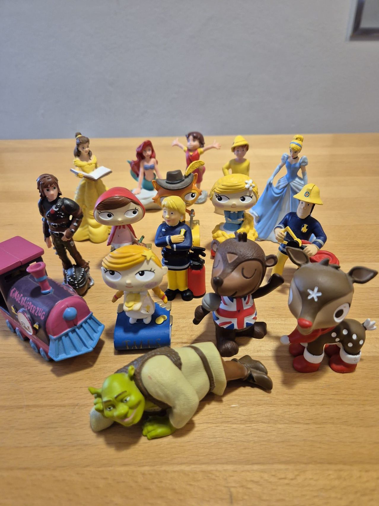 Several animated character figurines on a wooden surface. Includes characters like Belle, Cinderella, Ariel, Red Riding Hood, and Shrek. A train, a bear, and a reindeer are also present.