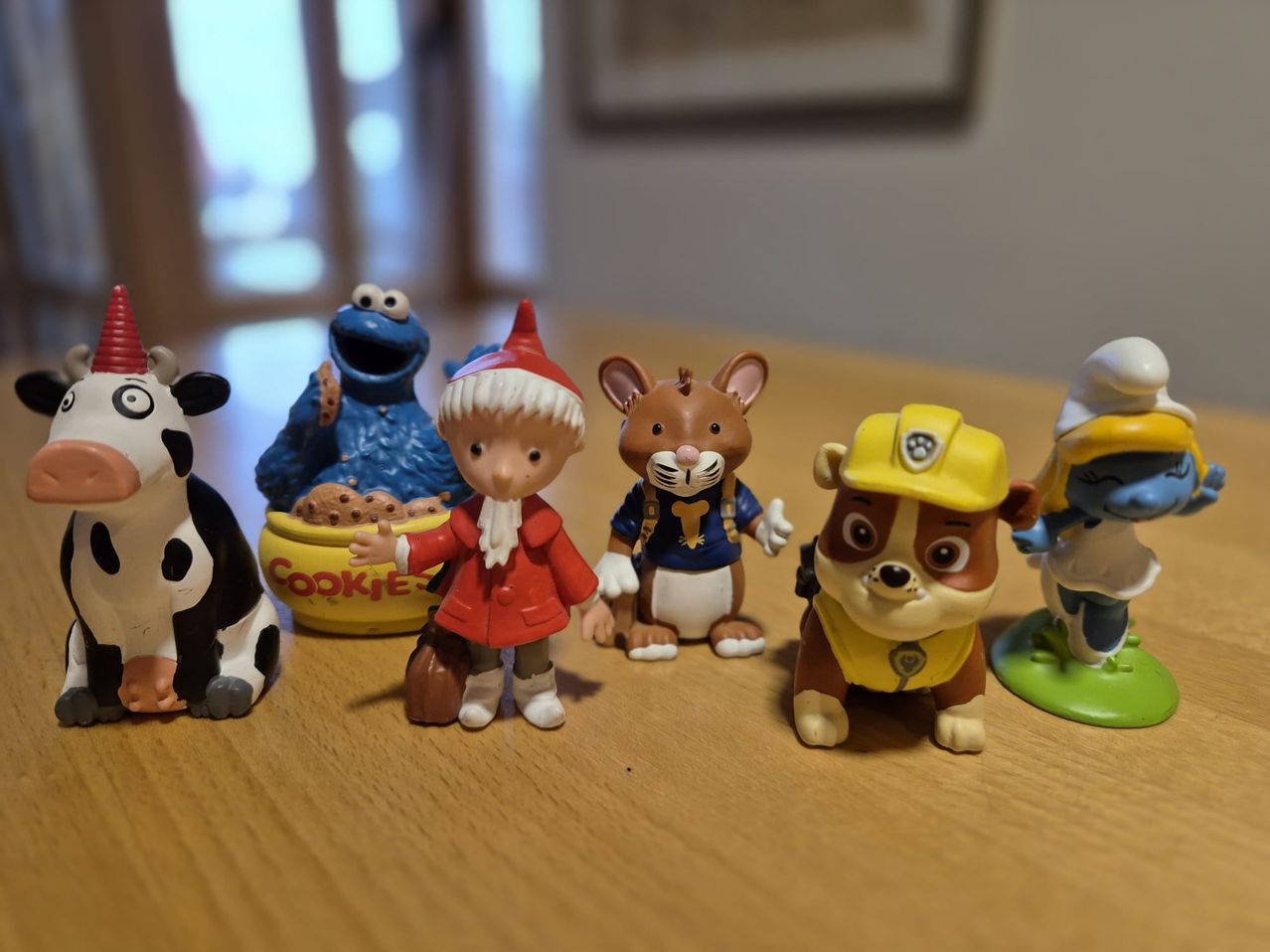 A collection of small figurines is on a wooden table. These include Cookie Monster, a Santa figure, a mouse, a dog, and a cow. The figurines are colorful and appear to be from different children's shows.