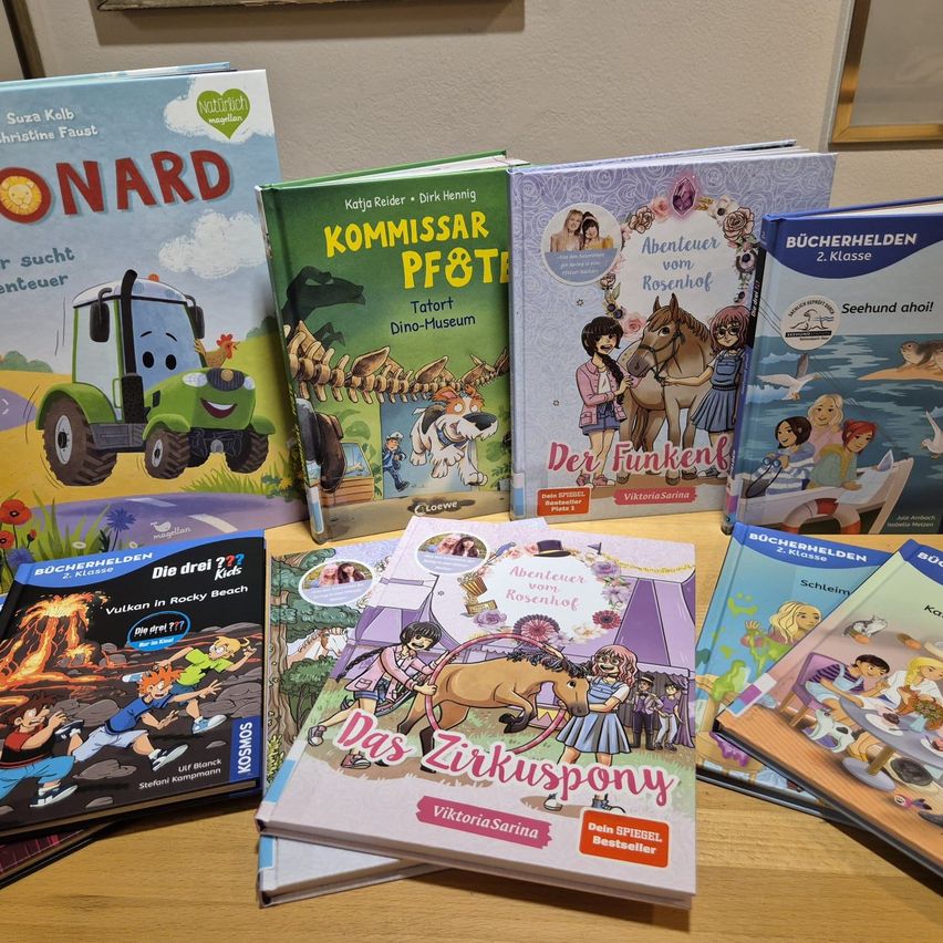A collection of children's books on a wooden table. Titles include 'Kommissar Pf&Tatort Dino-Museum,' 'Abenteuer vom Rosenhof,' and 'Das Zirkuspony.' There are also books titled 'Bücherhelden' and 'Der Funken.' The books are arranged in a row.