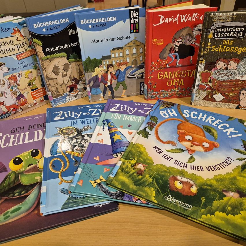 A collection of colorful children's books displayed on a wooden table, including titles like 'Die drei', 'Gangsta Oma', and 'Oh Schreck!'. Each book has a distinct cover design with characters and themes that appeal to children.