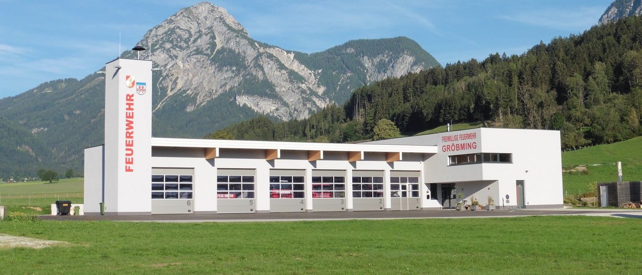 Ein weisses Feuerwehrhaus namens Freiwillige Feuerwehr Grobming steht vor einem grossen, grasbewachsenen Feld mit Bergen im Hintergrund.