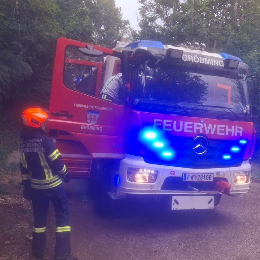 Ein Feuerwehrauto aus Grobming mit offener Fronttür ist auf einer Straße geparkt. Ein Feuerwehrmann steht daneben und trägt einen Helm und eine Uniform.