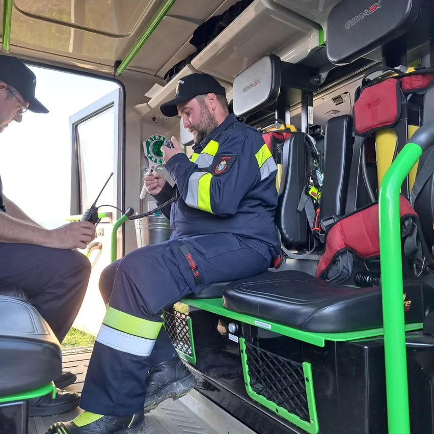 Zwei Männer sitzen in einem Fahrzeug. Einer hält ein Funkgerät und scheint dem anderen Mann etwas zu erklären. Das Innere des Fahrzeugs hat mehrere Sitze, Ablagen und einen grünen Handgriff.