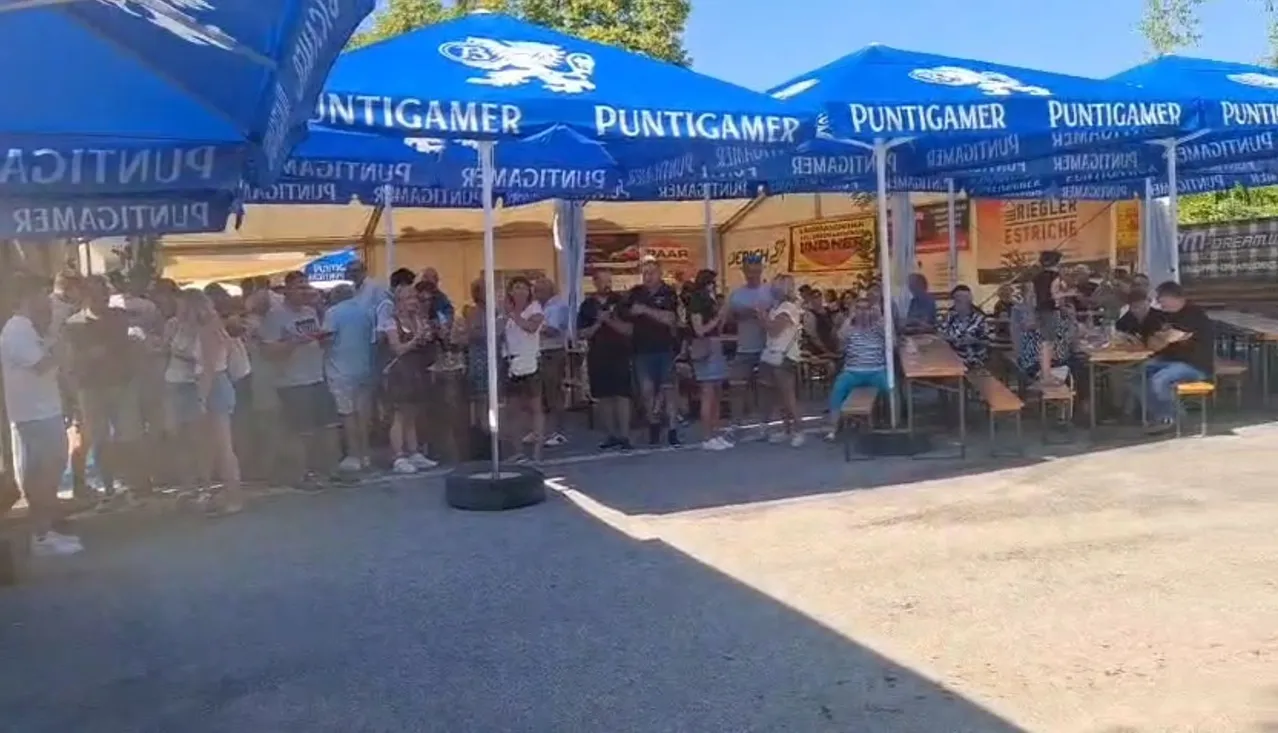 Eine Gruppe von Menschen steht unter blauen Zelten bei einer Veranstaltung im Freien, eine Person sitzt auf einer Bank und andere unterhalten sich und gehen herum.