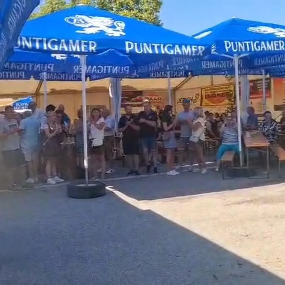 Eine Gruppe von Menschen steht unter blauen Zelten bei einer Veranstaltung im Freien, eine Person sitzt auf einer Bank und andere unterhalten sich und gehen herum.