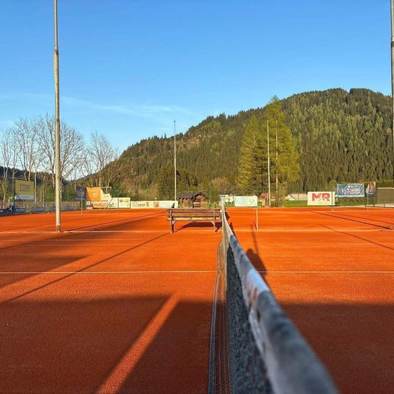 Ein leerer Tennisplatz mit einer Holzbank an der Seite. Ein Zaun steht vor dem Platz. Der Platz hat eine orange Oberfläche. Ein Gebirgszug liegt hinter dem Platz.