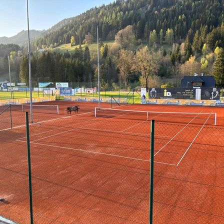 Ein leerer roter Sandtennisplatz mit einer Bank, umgeben von einem Zaun. Ein Berg mit Bäumen und einem Haus ist im Hintergrund.