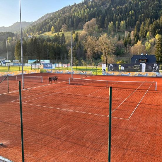 Ein leerer roter Sandtennisplatz mit einer Bank, umgeben von einem Zaun. Ein Berg mit Bäumen und einem Haus ist im Hintergrund.