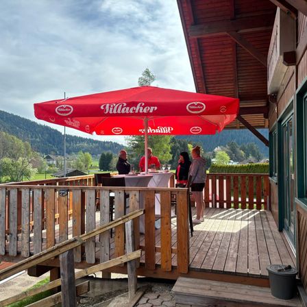 Eine Gruppe von Menschen versammelt sich unter einem roten Schirm auf einer Holzterrasse mit Blick auf eine Berglandschaft.