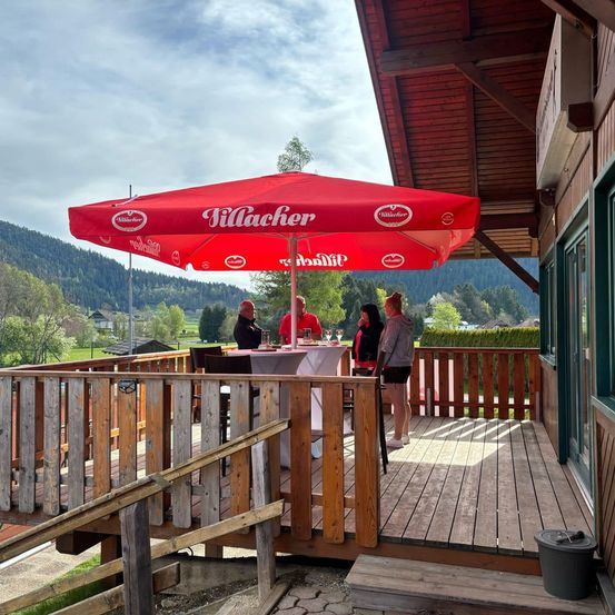 Eine Gruppe von Menschen versammelt sich unter einem roten Schirm auf einer Holzterrasse mit Blick auf eine Berglandschaft.