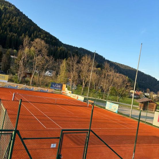 Ein leerer Tennisplatz mit roter Oberfläche, umgeben von einem grünen Zaun. Bäume säumen die Ränder, mit einem Berg und Häusern im Hintergrund. Ein klarer blauer Himmel ist über uns.