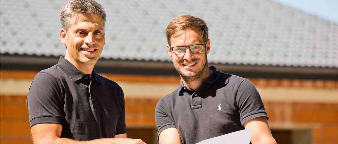 Zwei Männer in schwarzen Polo-Shirts stehen draußen, einer hält ein Papier in der Hand, sie scheinen etwas zu besprechen. Hinter ihnen befindet sich ein Gebäude mit einem grauen Dach.
