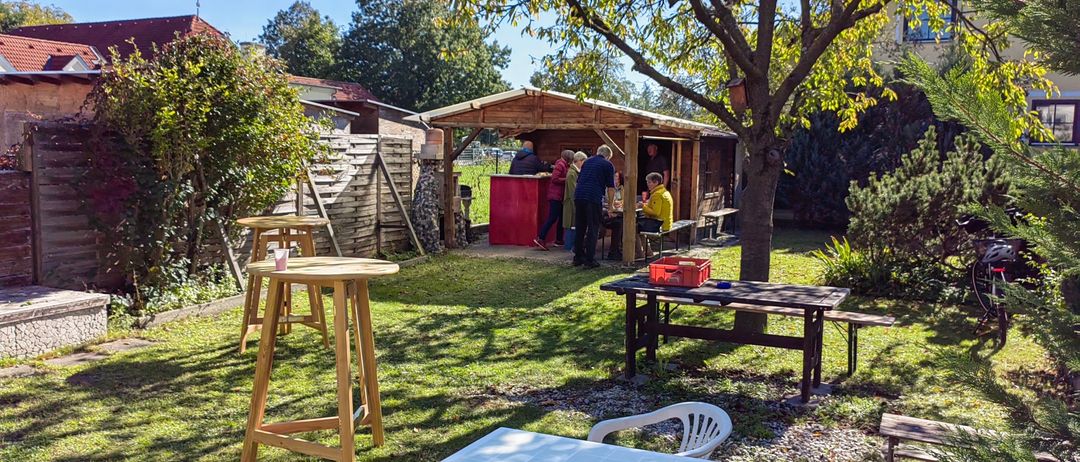 Freisitz mit einem hölzernen Unterstand. Mehrere Personen stehen um einen Tisch und eine rote Theke. Bäume und Bänke im Hintergrund.