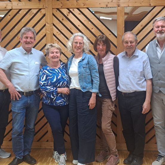 Eine Gruppe älterer Menschen posiert für ein Foto in einem Raum mit einer Holzwand. Sie alle lächeln und scheinen für eine Kamera zu posieren.
