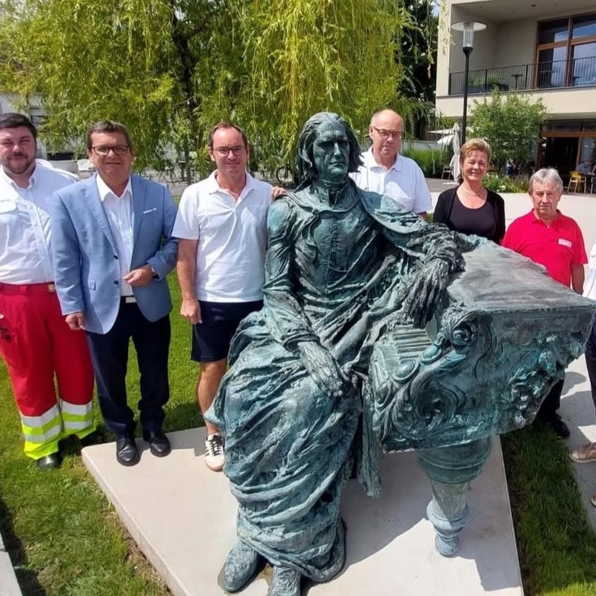 Eine Gruppe von Menschen, schick gekleidet, steht um eine große Bronzestatue eines Mannes, der auf einer Bank sitzt, im Freien vor einem Gebäude.
