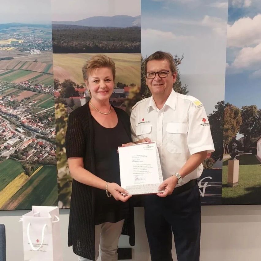 Ein Mann und eine Frau stehen lächelnd und halten ein Zertifikat vor einer malerischen Kulisse. Der Mann trägt ein weißes Uniform mit einem roten Kreuz, während die Frau in Freizeitkleidung ist.