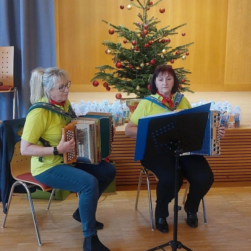 Zwei Frauen sitzen auf Stühlen und spielen Akkordeons, möglicherweise in einer Kirche oder einem Veranstaltungssaal. Eine schaut die andere an. Hinter ihnen steht ein Weihnachtsbaum mit Geschenken und einem blauen Vorhang.