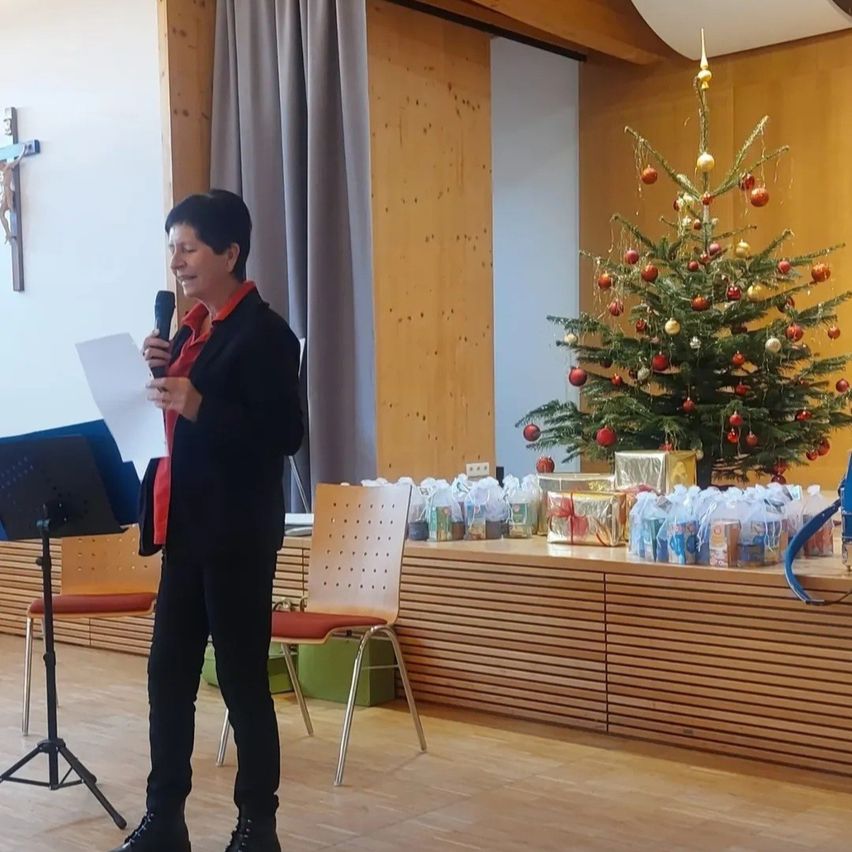 Eine Frau in Schwarz hält ein Mikrofon und Papier und steht vor einem geschmückten Weihnachtsbaum und einer Bühne. Hinter ihr befindet sich ein Kreuz an der Wand.