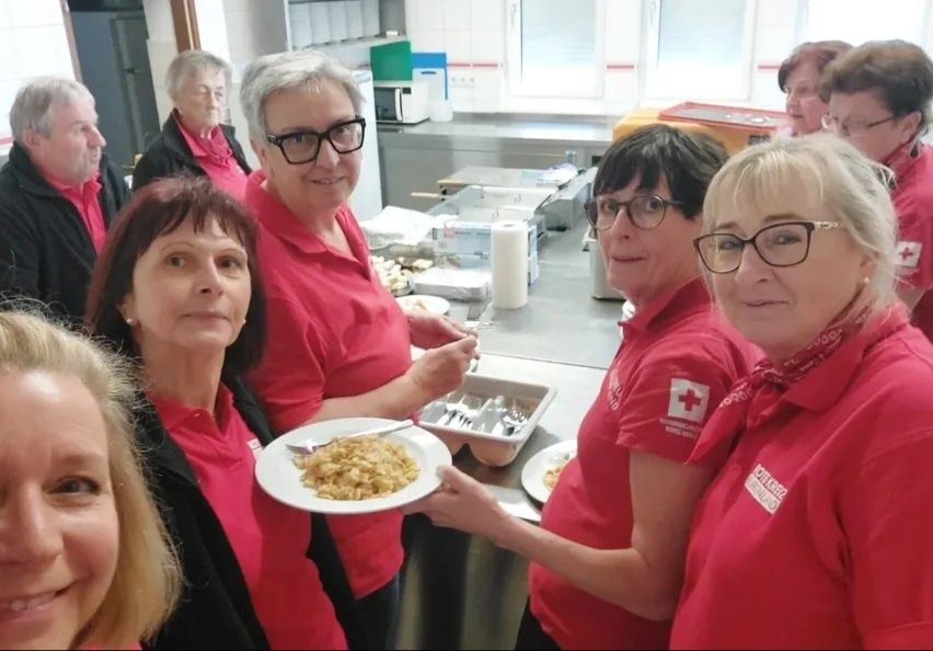 Eine Gruppe von Frauen in roten Shirts steht in einer Küche. Sie lächeln und halten einen Teller mit Essen. Ein rotes Kreuz-Logo ist auf ihren Shirts sichtbar.