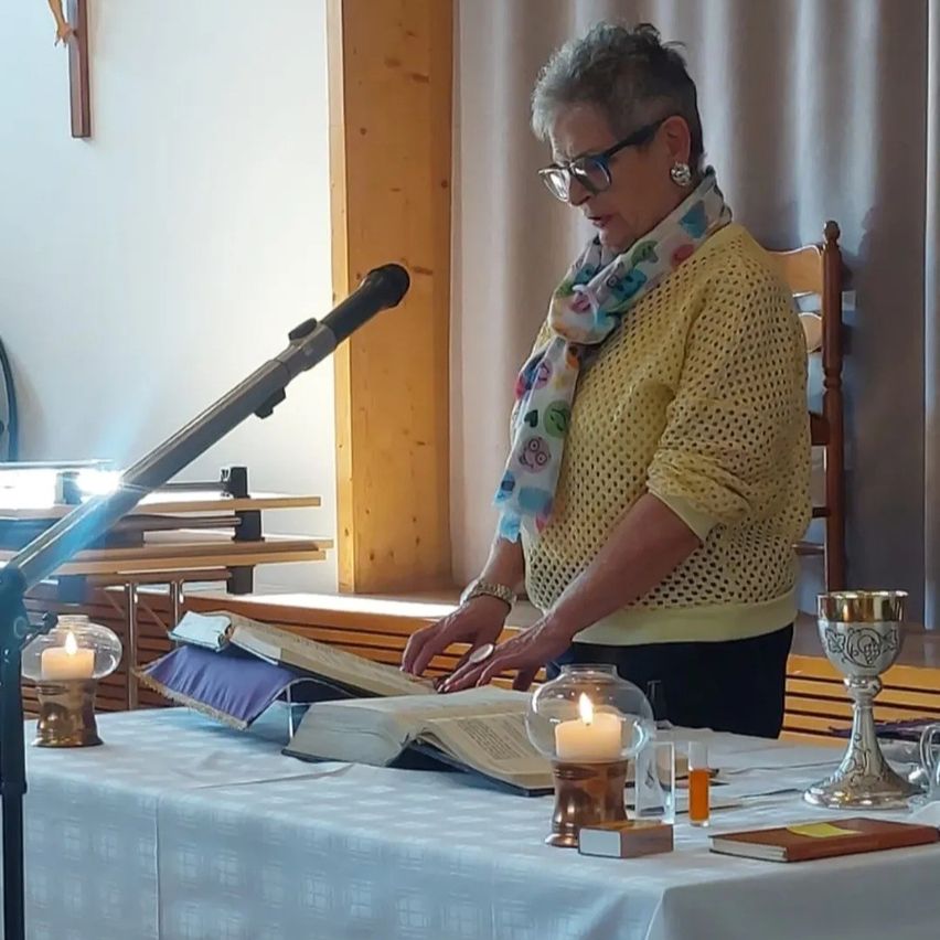Eine ältere Frau mit Brille und Schal steht an einem Tisch und liest aus einem Buch vor.