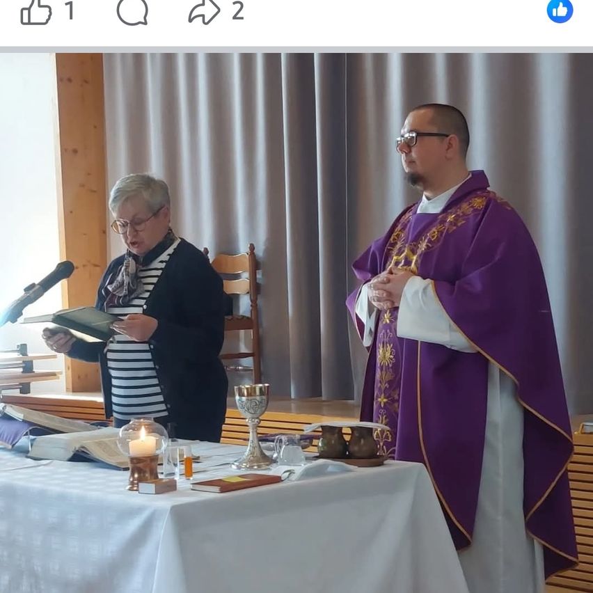 Ein Priester steht am Altar in einer Kirche mit einer Frau, die ein Buch liest, und einer Kerze auf dem Tisch.