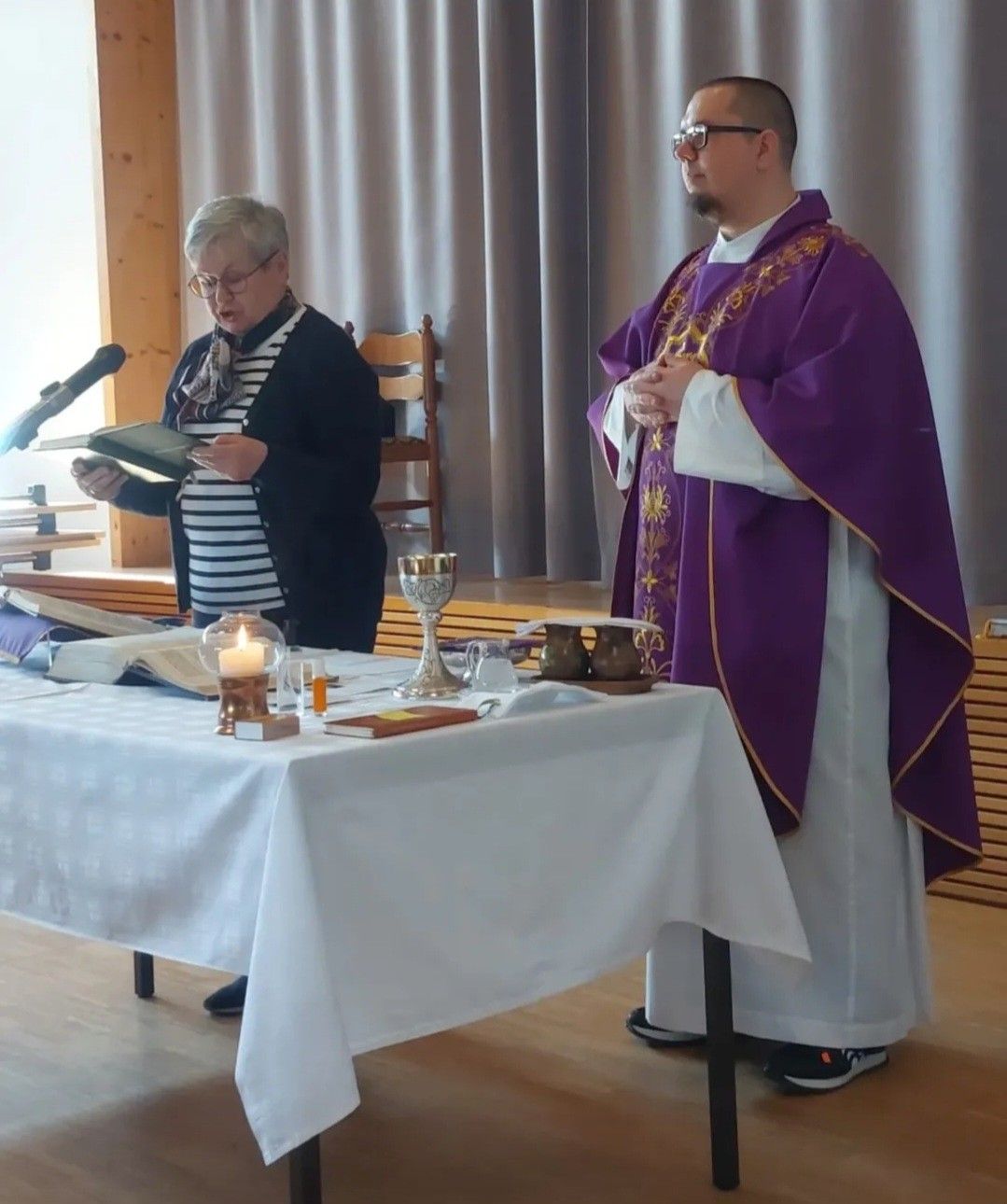 Ein Priester und eine Frau stehen in einer Kirche und lesen aus einem Buch. Vor ihnen steht ein Tisch mit religiösen Gegenständen.