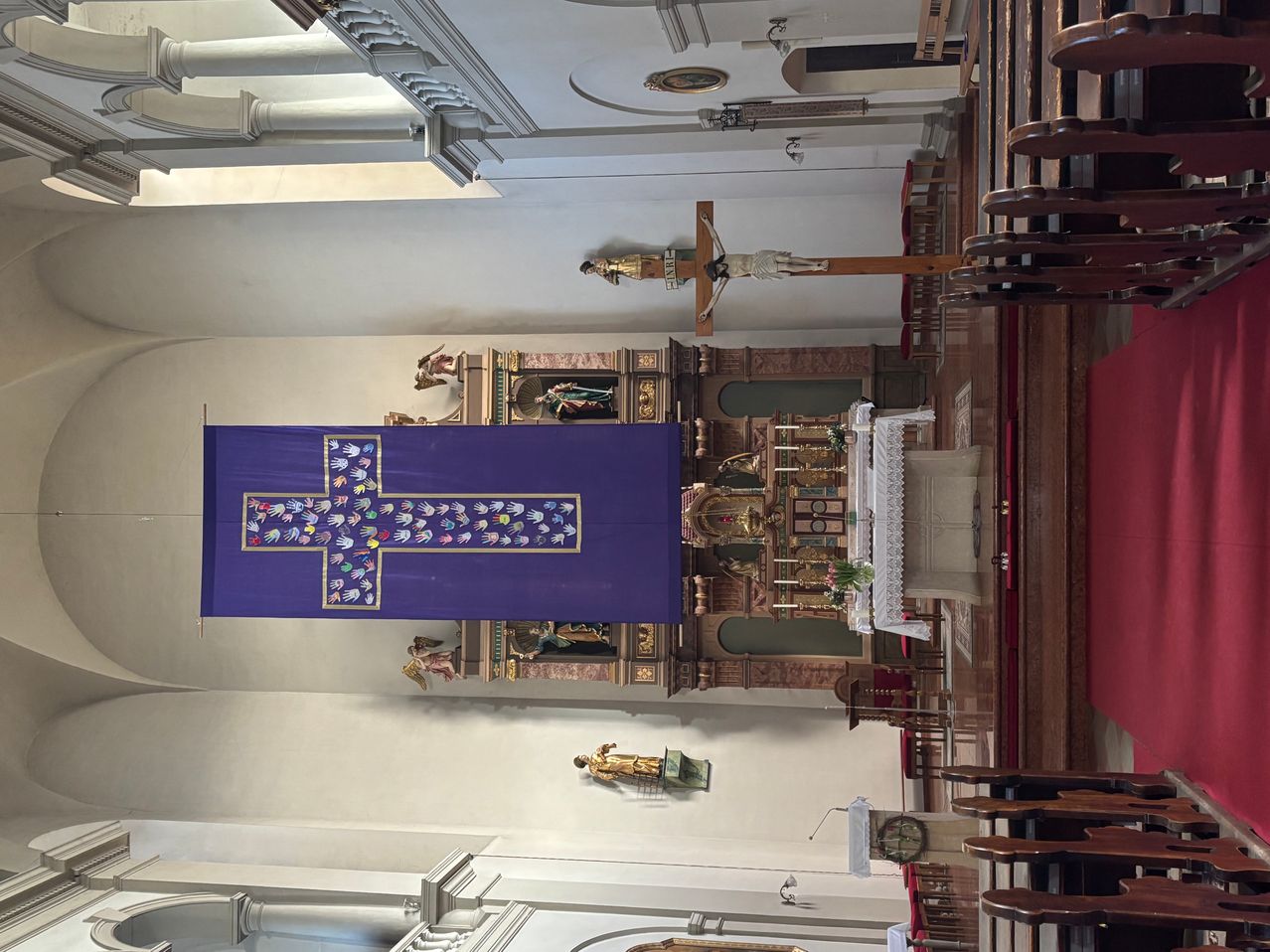 A church interior with a purple banner featuring a colorful cross. In the background, a crucifix hangs above an altar with a statue. Pews line the sides.