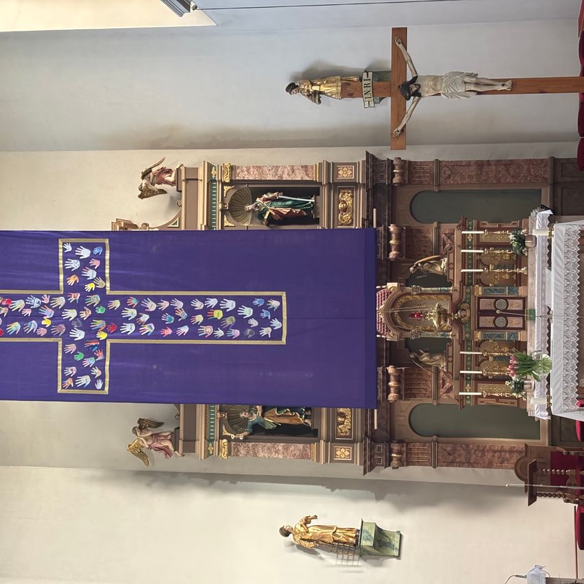 An aerial view of a church altar adorned with a large purple banner featuring a golden cross. Multiple statues and religious symbols are present.
